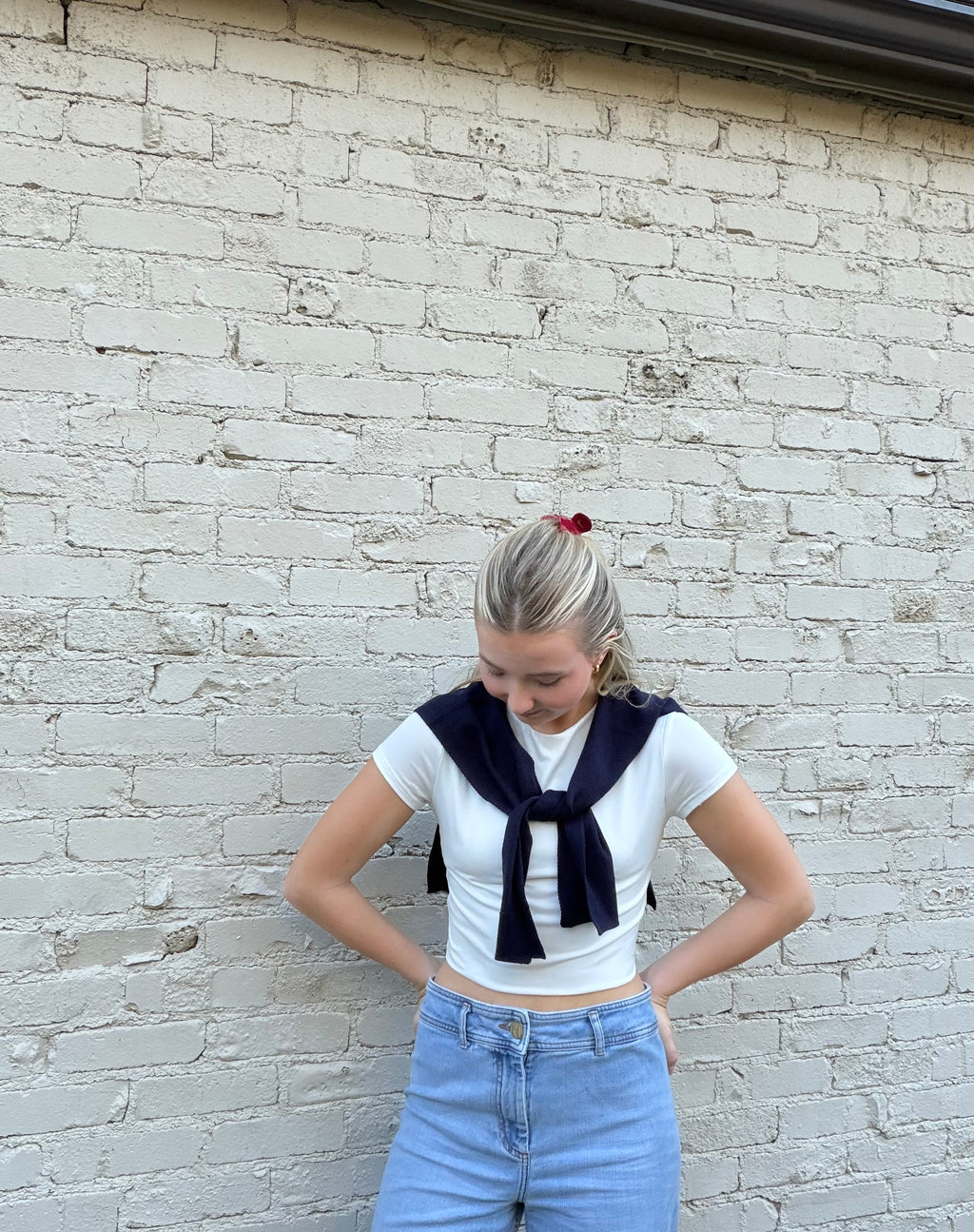 A young woman with blonde hair tied back stands against a white painted brick wall, wearing a white short-sleeve crop top with a navy blue necktie detail, light blue jeans, and a small red hair accessory.