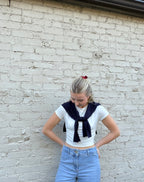 A young woman with blonde hair tied back stands against a white painted brick wall, wearing a white short-sleeve crop top with a navy blue necktie detail, light blue jeans, and a small red hair accessory.