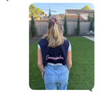 A woman viewed from behind wearing a navy blue sleeveless vest with the word 'Commandos' in white script on the back, paired with light blue jeans, standing on a green lawn in a backyard.